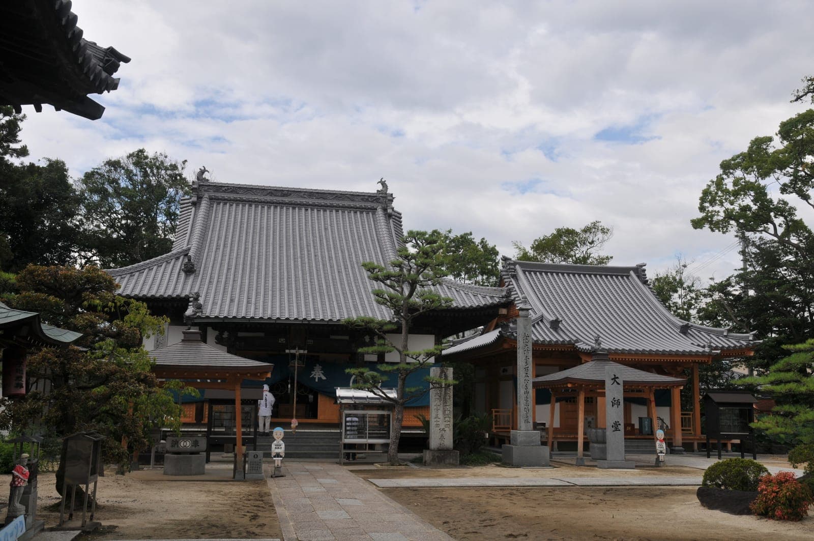 Esterno del tempio Sairin-ji con tetti di tegole, pilastri di pietra e alberi sotto un cielo nuvoloso.