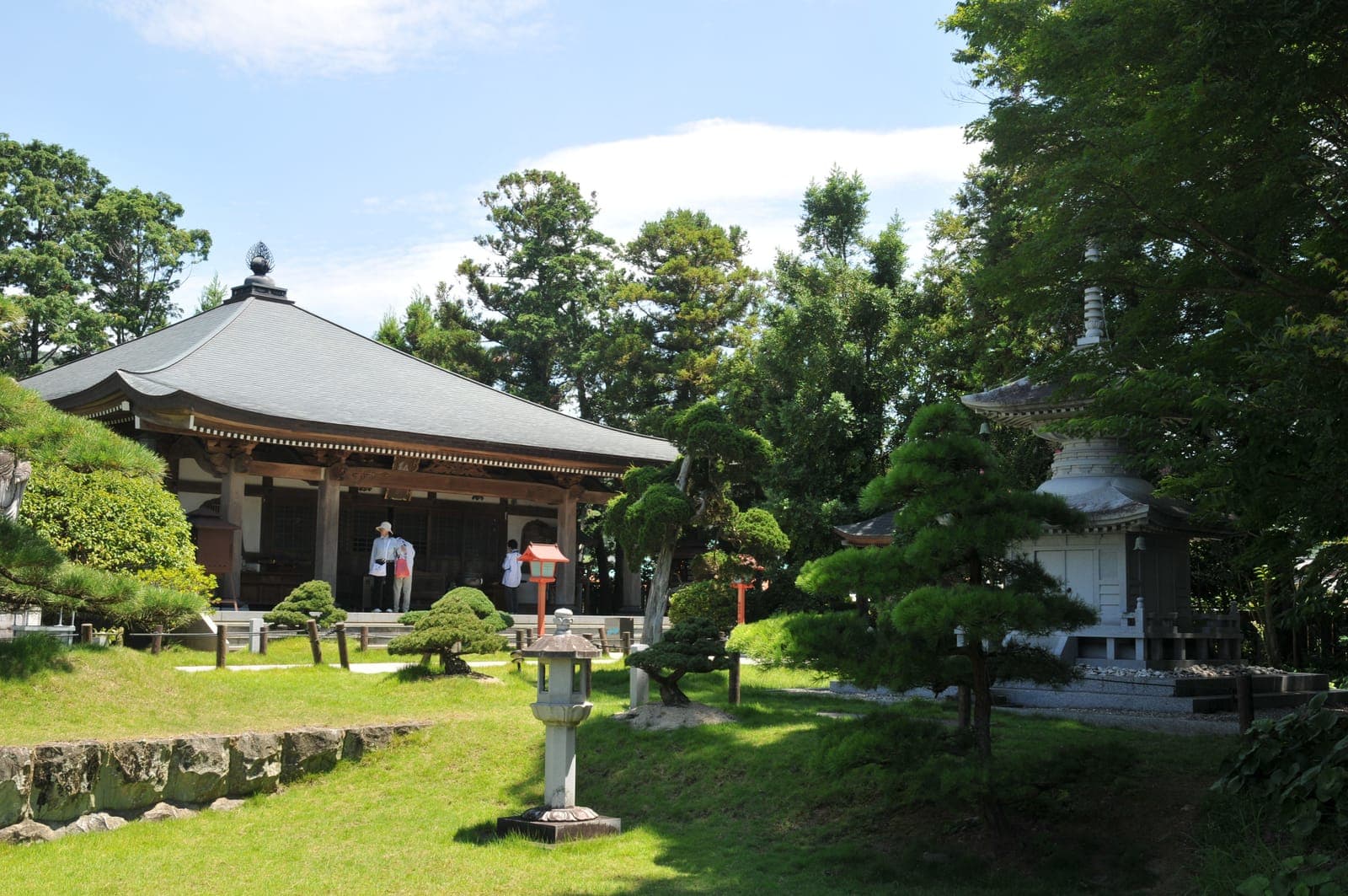 Kanjizai-ji temple grounds with a main hall, stone lanterns, trimmed trees, and a pagoda-like structure under a blue sky.