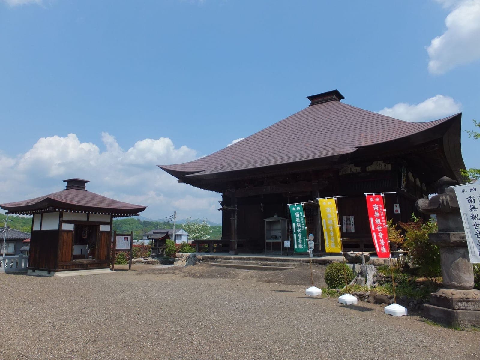 Exterior do templo Ryuseki-ji com grande salão de madeira, pequeno pavilhão, bandeiras e pátio de cascalho sob céu azul.