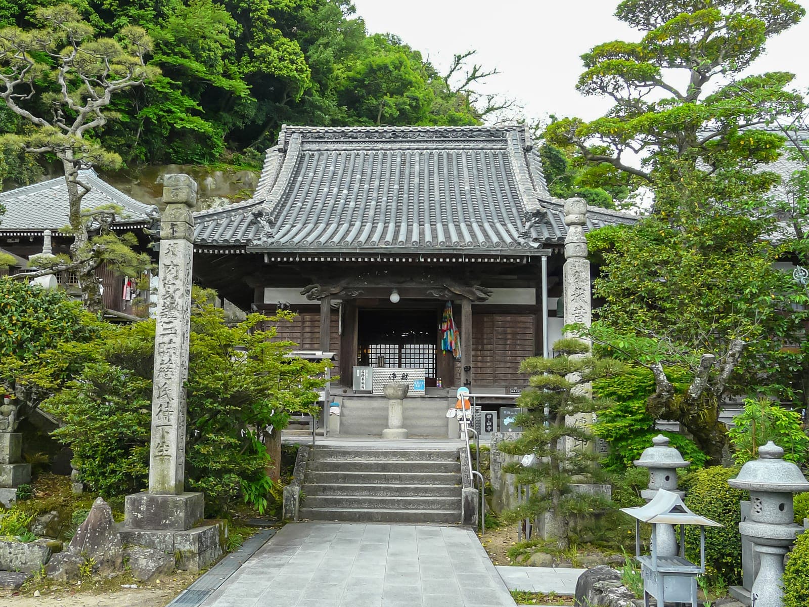 Exterior of Koyama-ji temple with a tiled roof, stone pillars, steps, and dense green trees around the entrance.