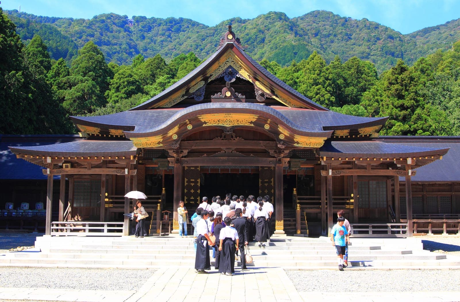 Edificio del santuario Iyahiko Jinja con tetto curvo decorato, persone radunate all’ingresso e montagne verdi sullo sfondo.