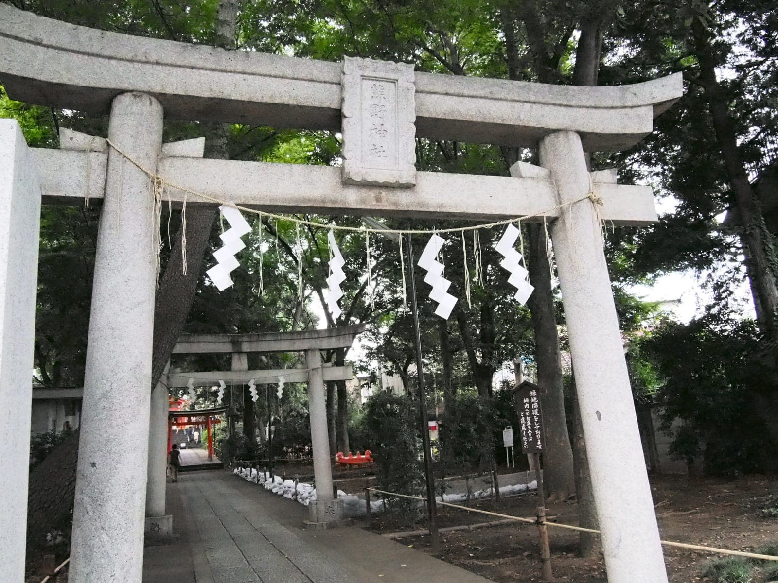 Portails torii en pierre à Jiyugaoka Kumano, avec shimenawa et bandes de papier le long d’un chemin bordé d’arbres.