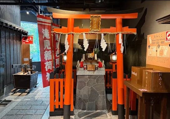 Santuário Takimi Inari com torii laranja vivo, corda sagrada e pequeno altar sob uma cobertura.
