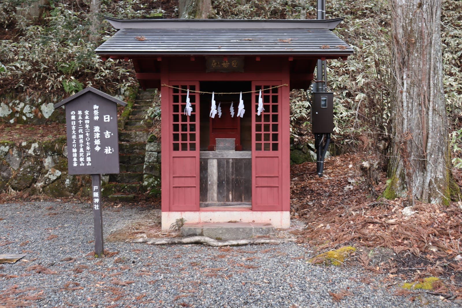Small red shrine building at Togakushi Jinja Chusha, with open doors, a hanging rope, and a signboard beside it.