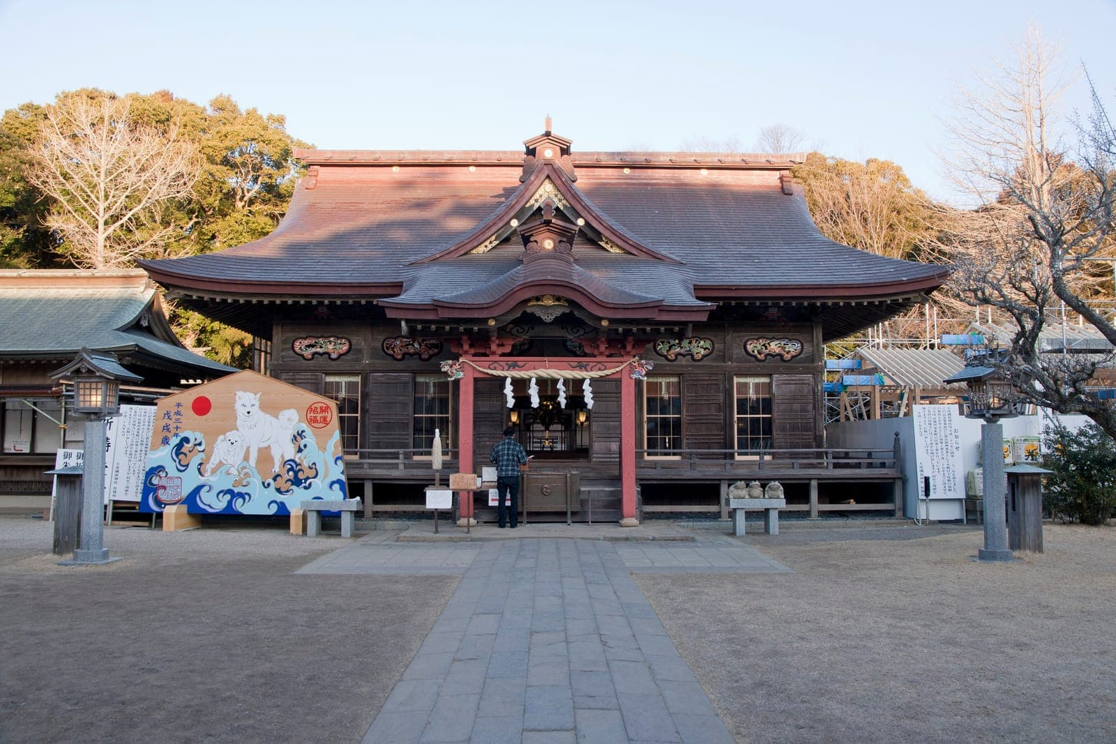 Exterior of Oarai Isosaki Jinja, showing a wooden shrine building with a curved roof, stone path, lanterns, and a person at the entrance.
