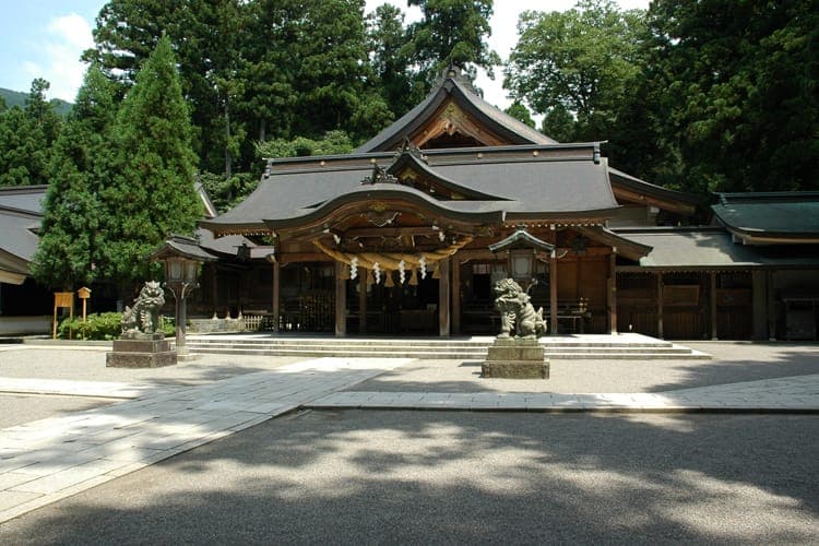 Exterior de Shirayama Hime Jinja, com um edifício de madeira, estátuas guardiãs de pedra e árvores altas ao fundo.
