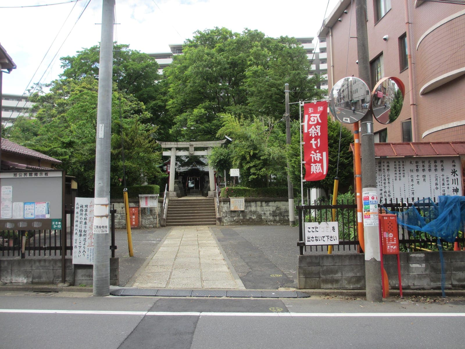 Entrada del templo Nakano Hikawa, con sendero de piedra, escalones y torii, rodeada de árboles, edificios urbanos y señales.