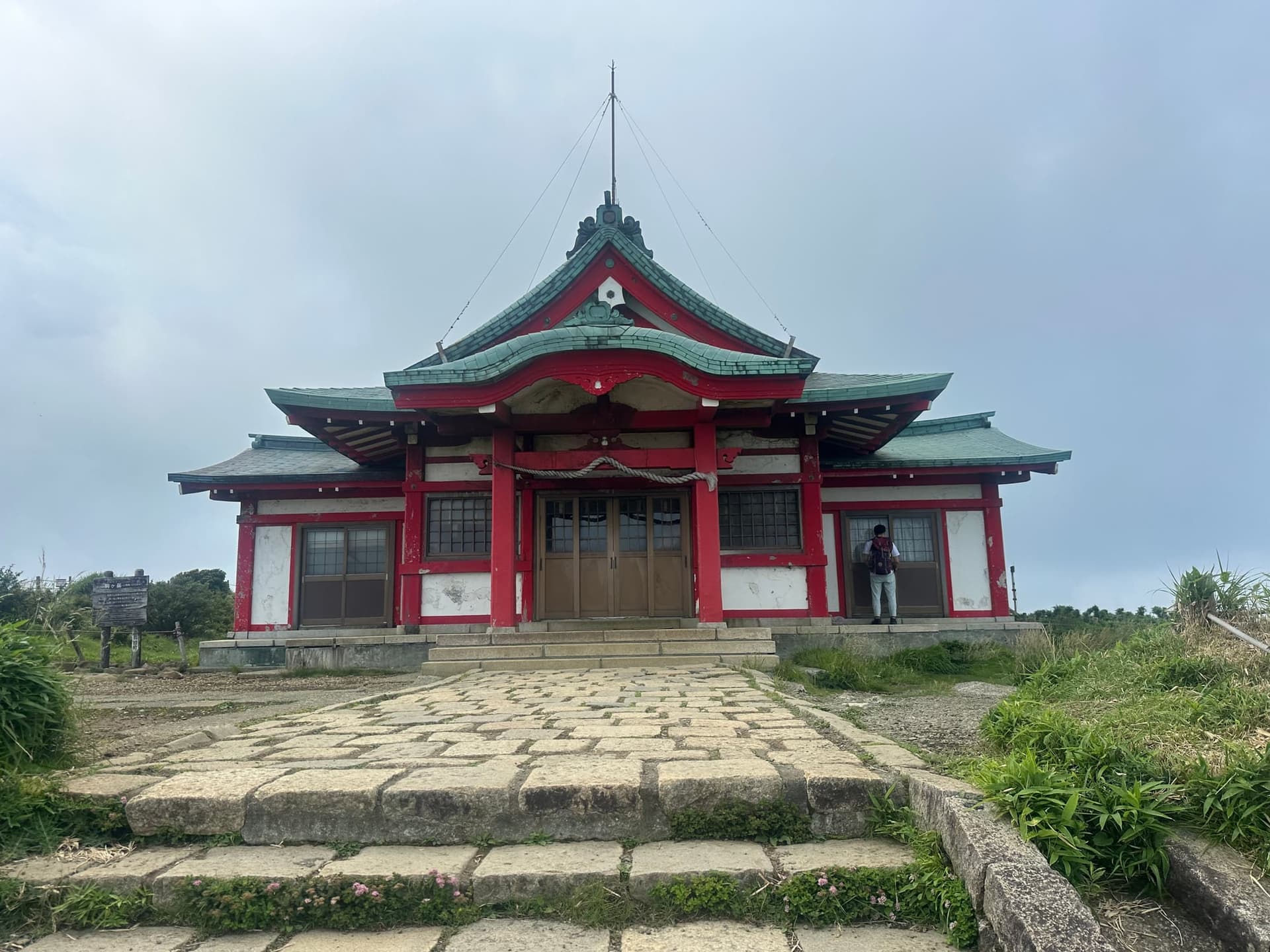 Exterior do templo Hakone Mototsumiya, com paredes vermelhas e brancas, telhado verde, degraus de pedra e uma pessoa à direita.