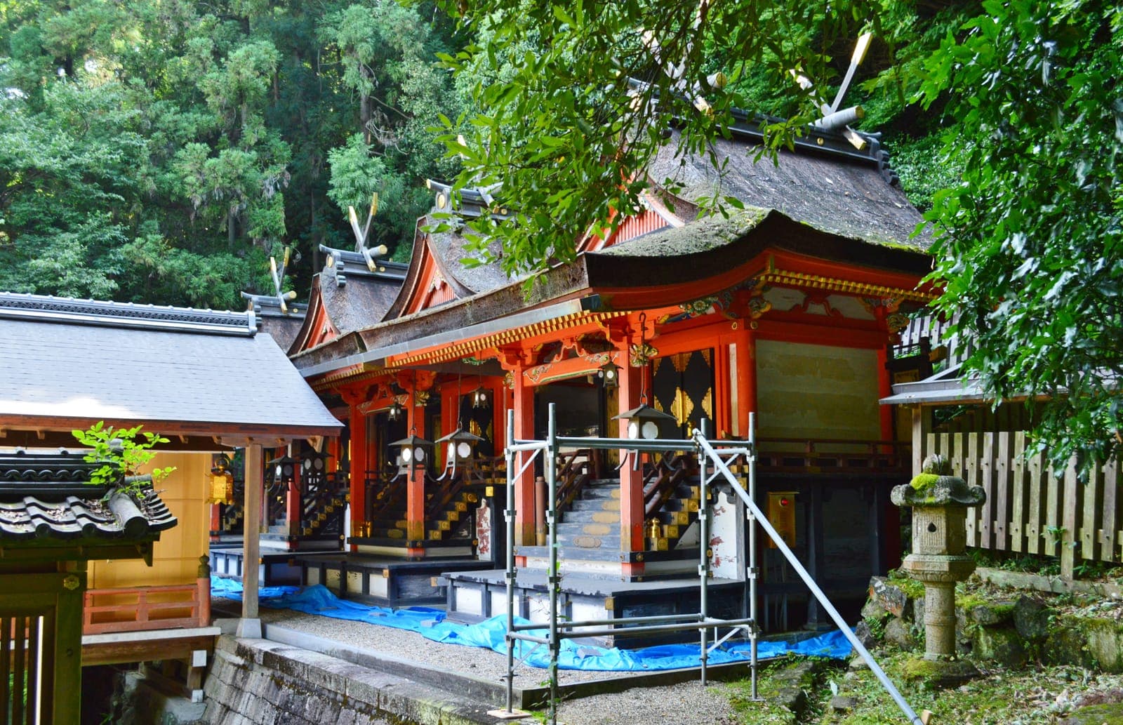 Edifícios do santuário Hiraoka Jinja com pilares vermelhos e telhados curvos, parcialmente cercados por andaimes e árvores.