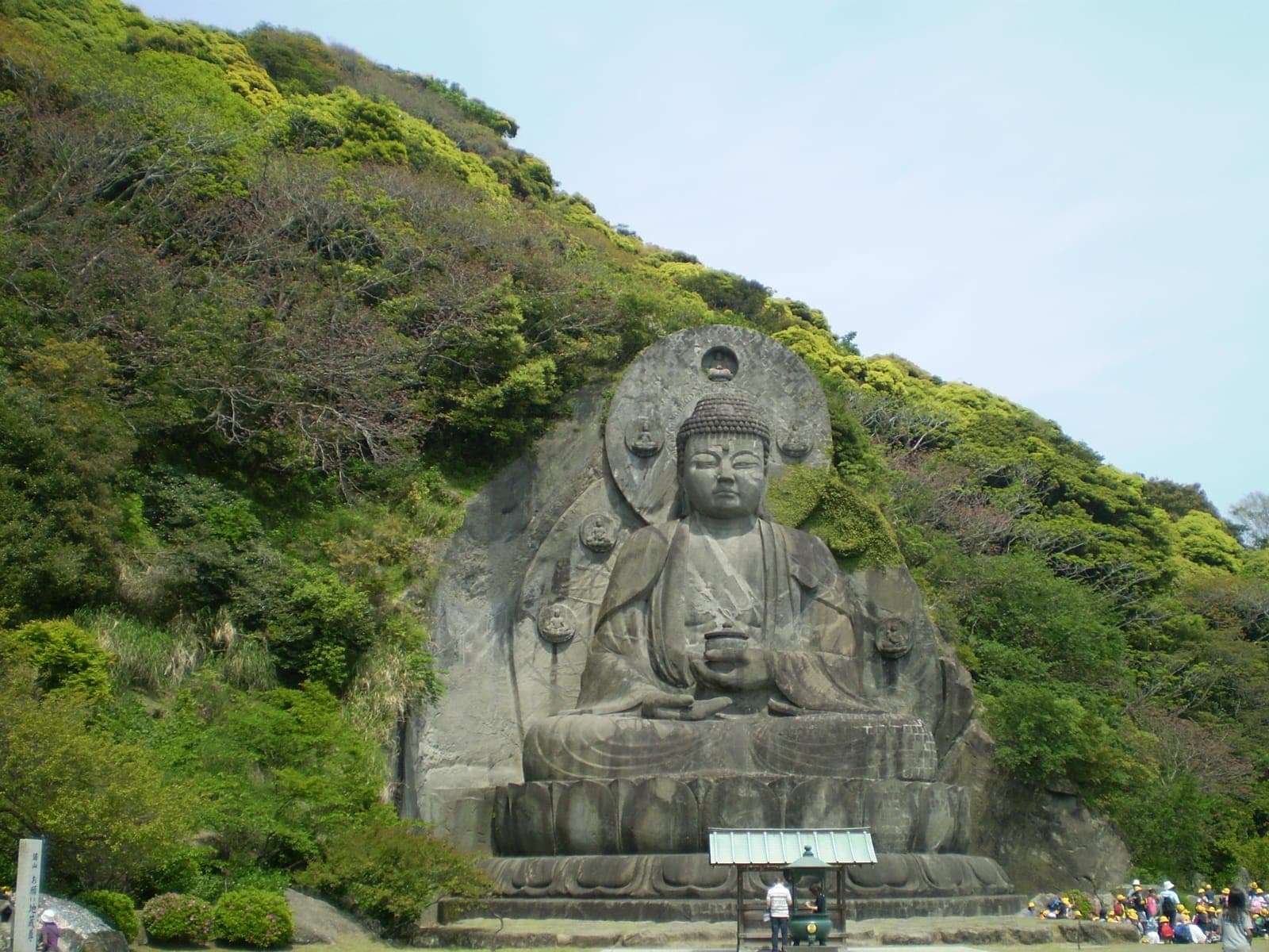Nihon-ji exterior with a large stone Buddha carved into a hillside, surrounded by green trees and visitors below.