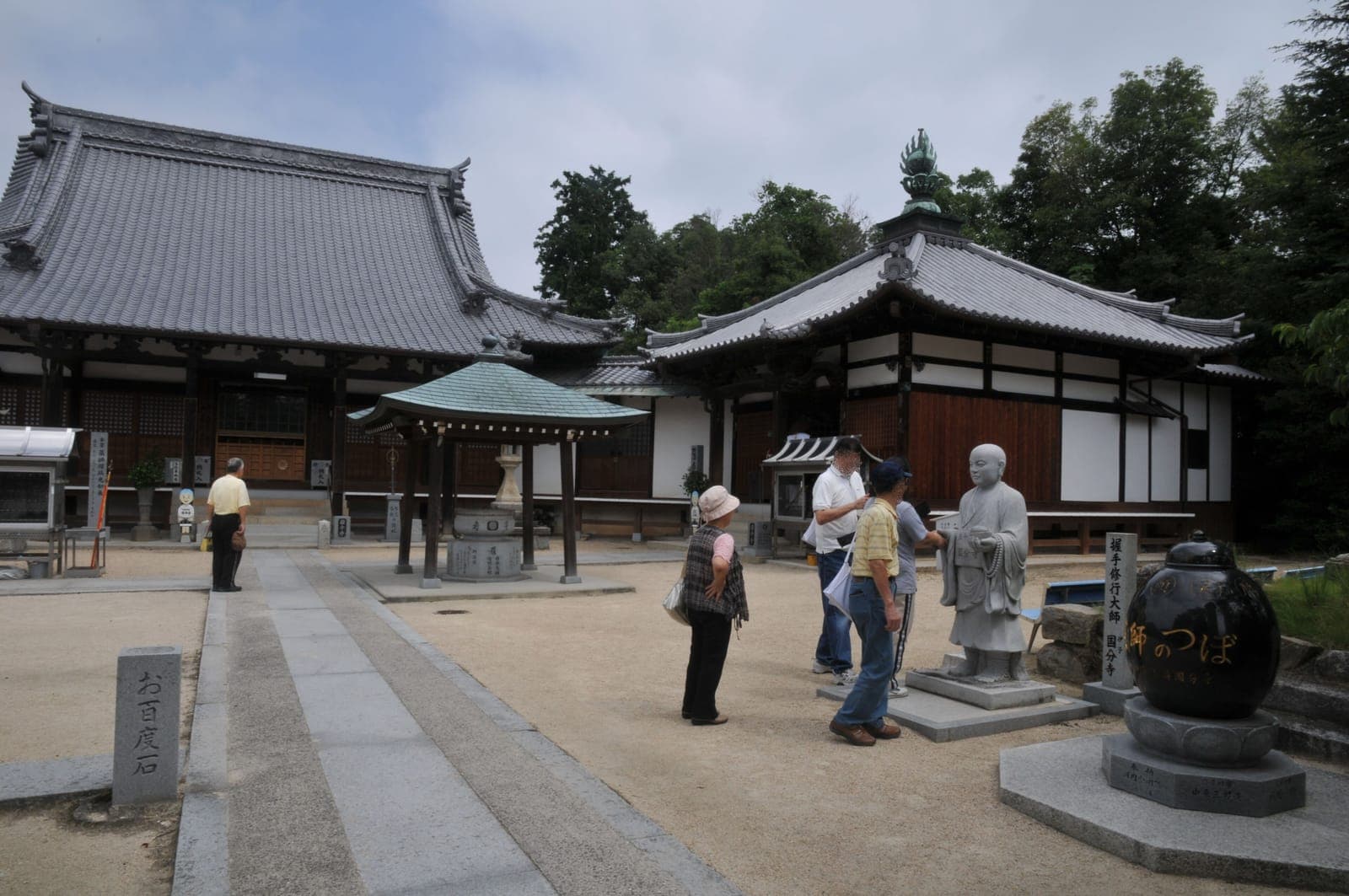Courtyard at Iyo Kokubun-ji with tiled temple buildings, a small pavilion, a stone statue, and several visitors.