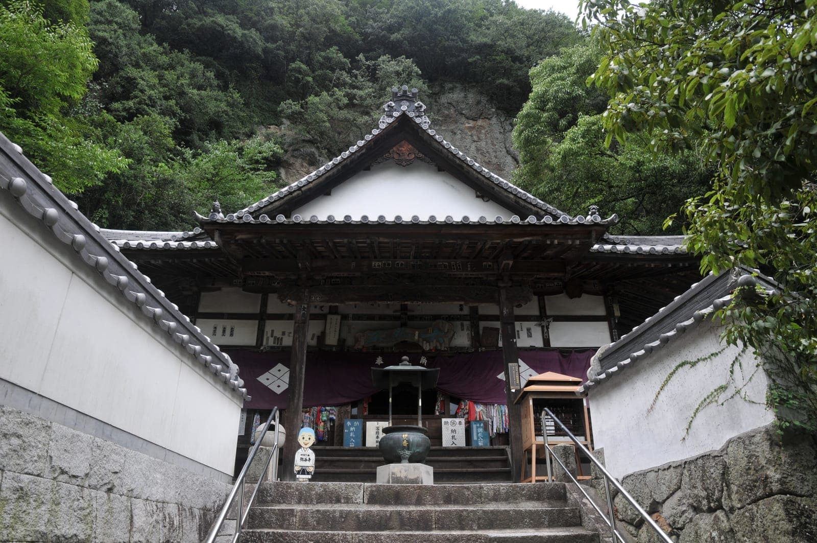 Iyadani-ji temple entrance with stone steps, white walls, tiled roof, and dense trees and cliff behind.