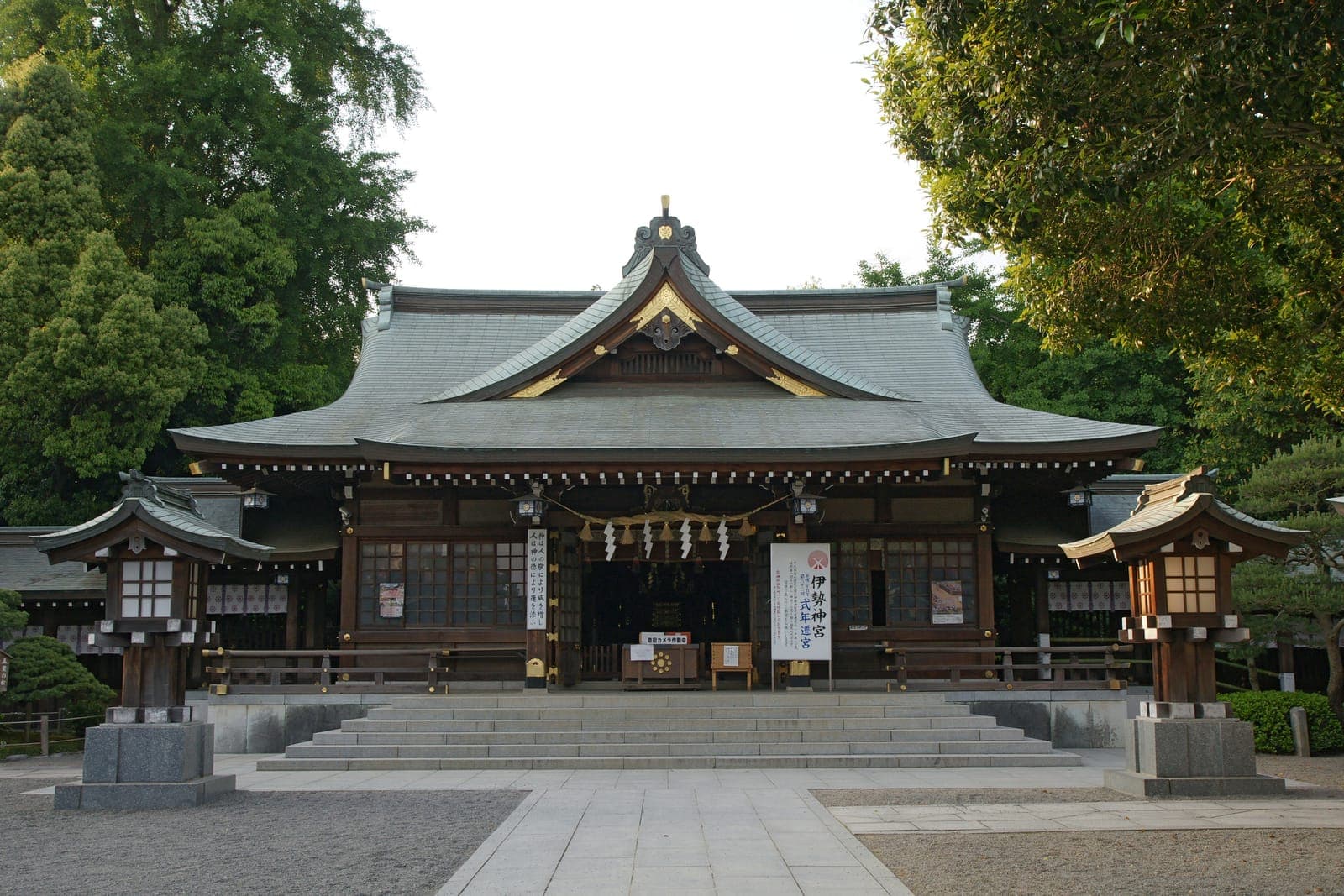 Extérieur du temple Izumi, avec large toit courbé, marches en pierre, lanternes et arbres autour de l’entrée.