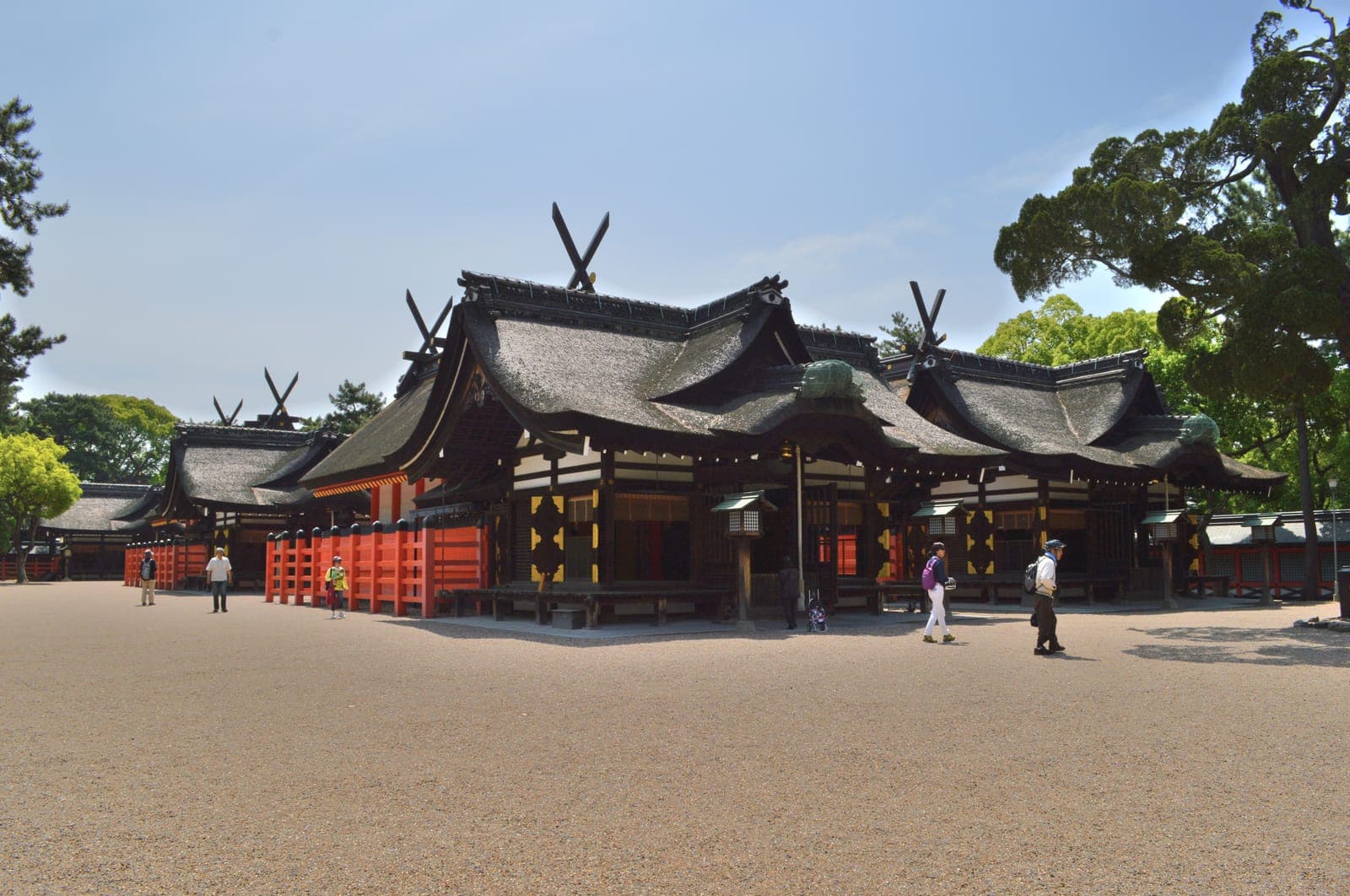 Esterno di Sumiyoshi Taisha con diversi edifici del santuario, tetti di paglia curvi, recinzioni rosse e alcuni visitatori nel cortile ghiaioso.