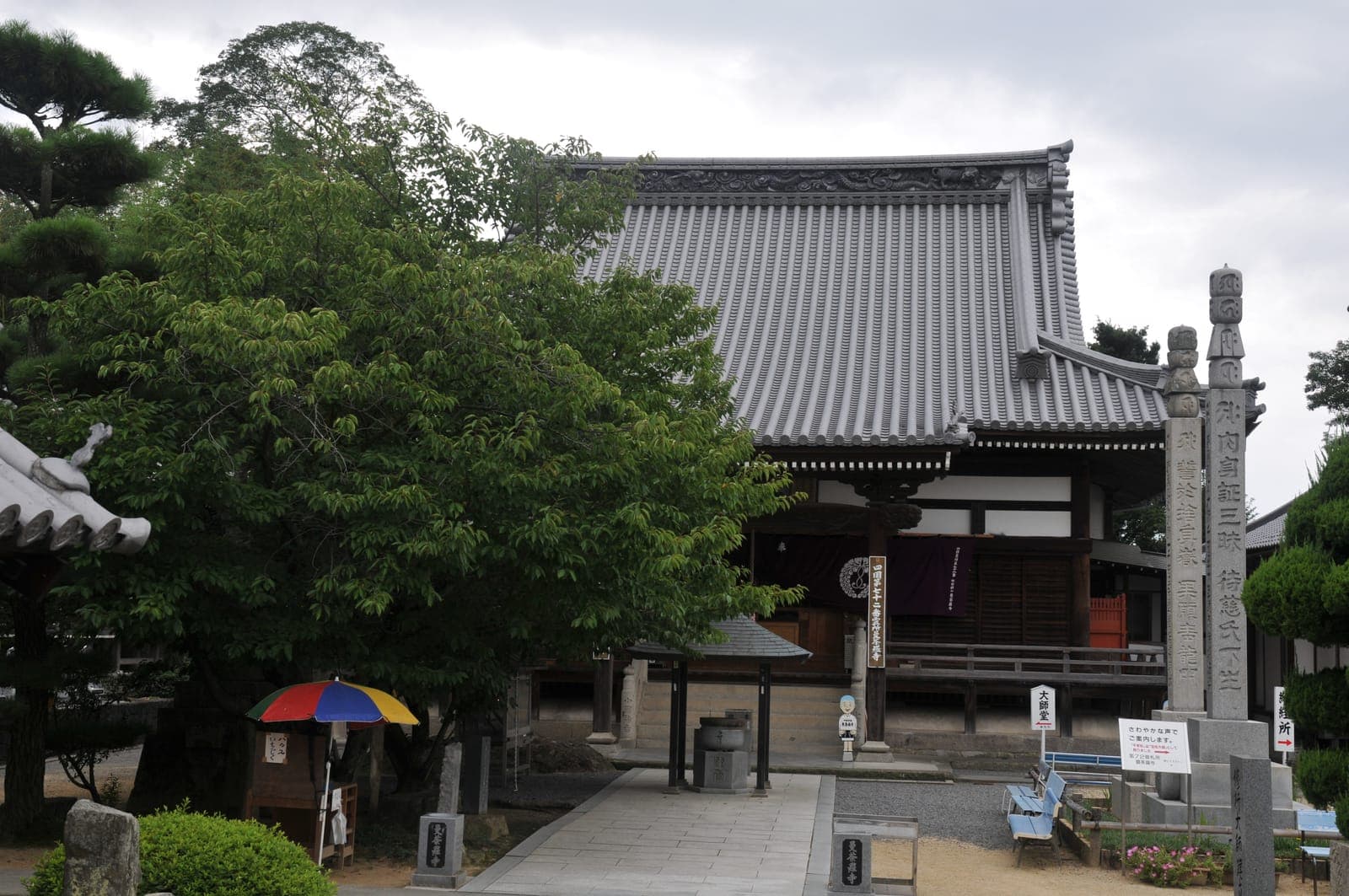 Mandara-ji temple exterior with a large tiled roof, stone pillars, and trees in the foreground.