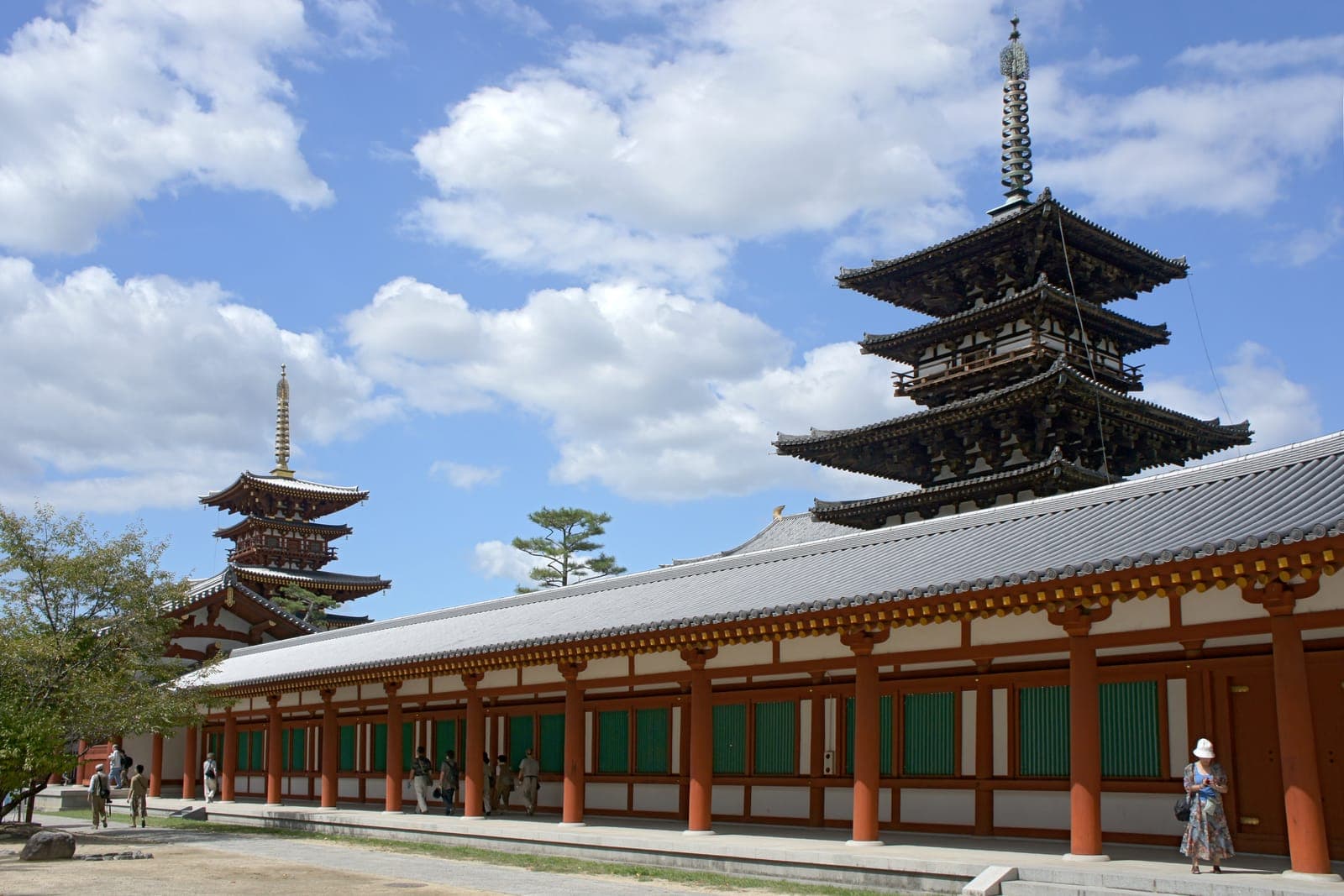 Esterno del tempio Yakushi-ji con due pagode, un lungo corridoio e visitatori sotto un cielo azzurro nuvoloso.
