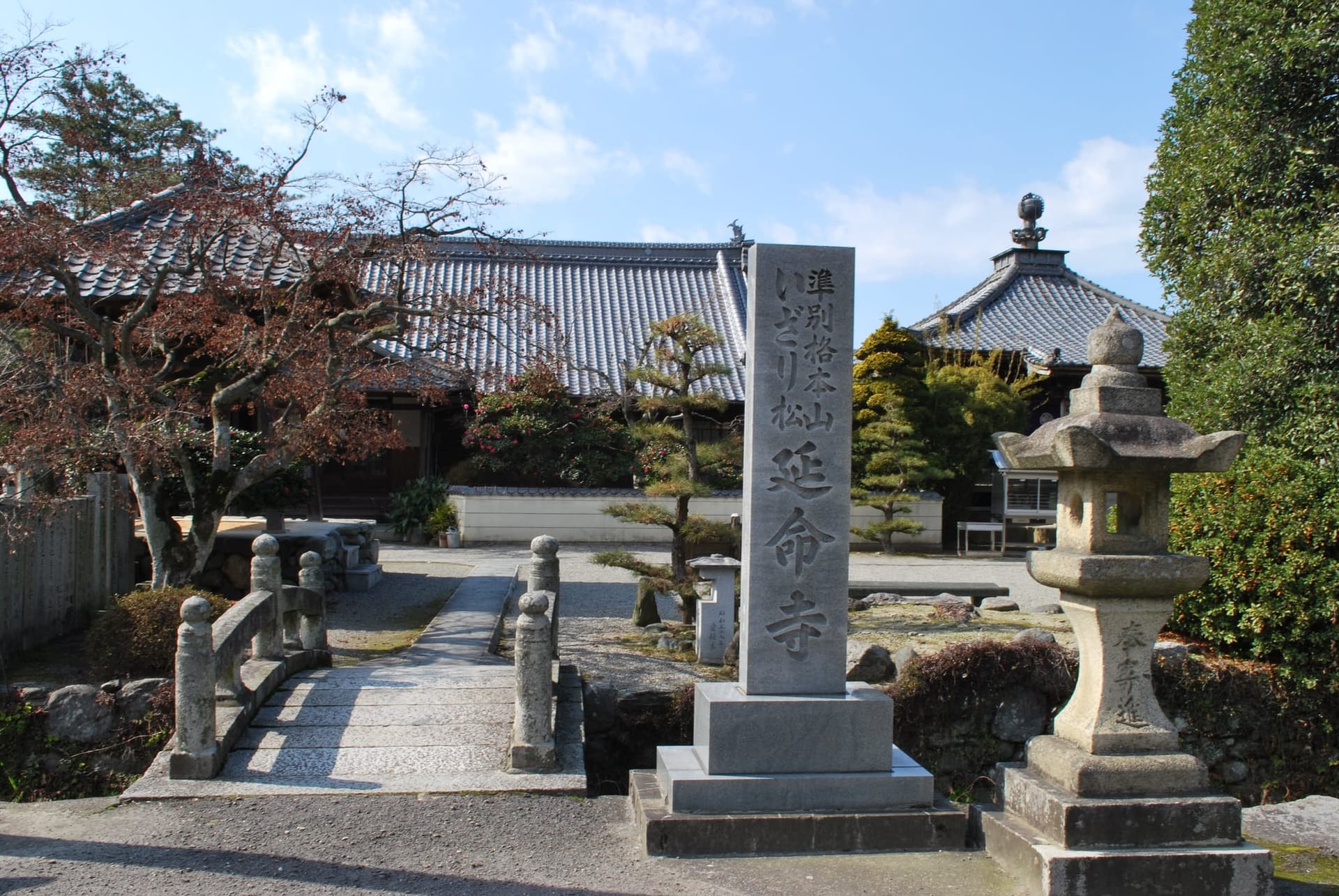 Ingresso del tempio Enmei-ji con ponte di pietra, lanterna in pietra e alta stele incisa davanti a edifici con tetti di tegole.