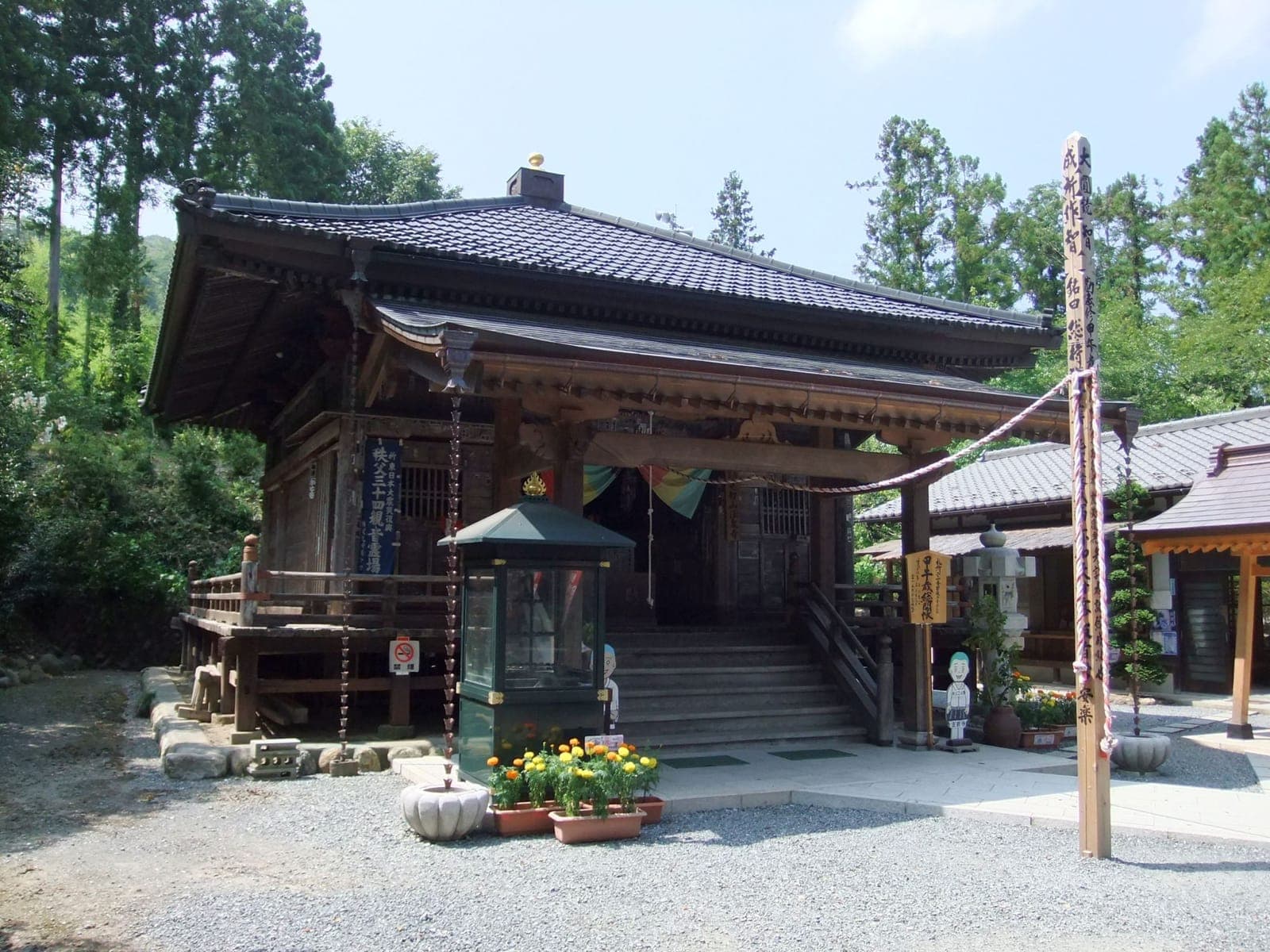 Exterior do templo Hosen-ji com salão de madeira, degraus, cordas pendentes, flores em vasos e árvores ao fundo.