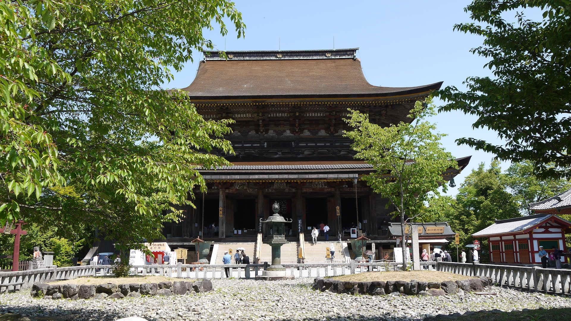 Exterior do templo Kinpusen-ji com grande salão de madeira, telhado curvo amplo, degraus de pedra e árvores em primeiro plano.