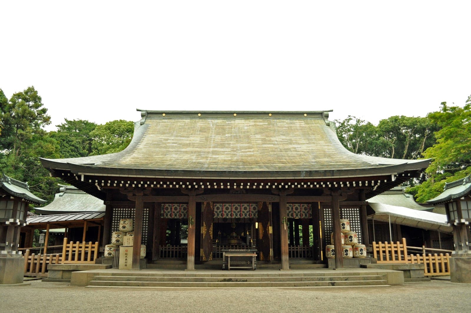 Gebäude des Musashi Ichinomiya Hikawa-Schreins mit breitem geschwungenem Dach, Stufen, Laternen und Bäumen dahinter.
