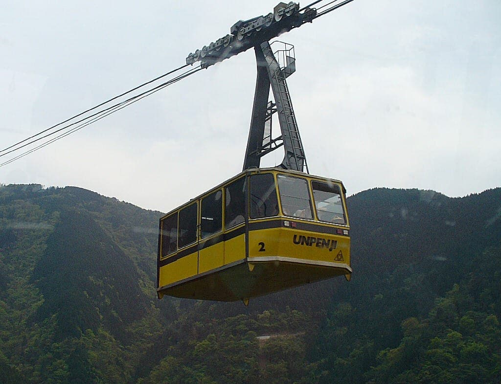 Téléphérique jaune d’Unpen-ji suspendu au-dessus de montagnes boisées sous un ciel nuageux.