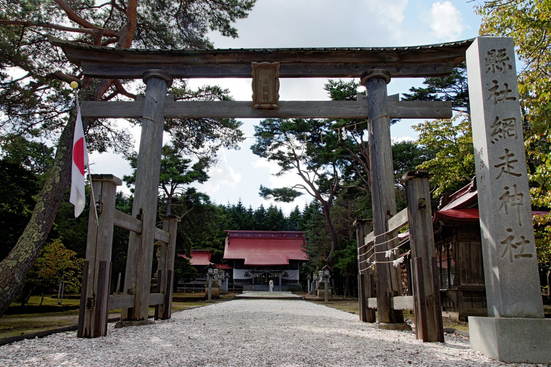 Ingresso di Abashiri Jinga con torii in legno, vialetto di ghiaia, pilastro di pietra, bandiera giapponese e edifici sul fondo.