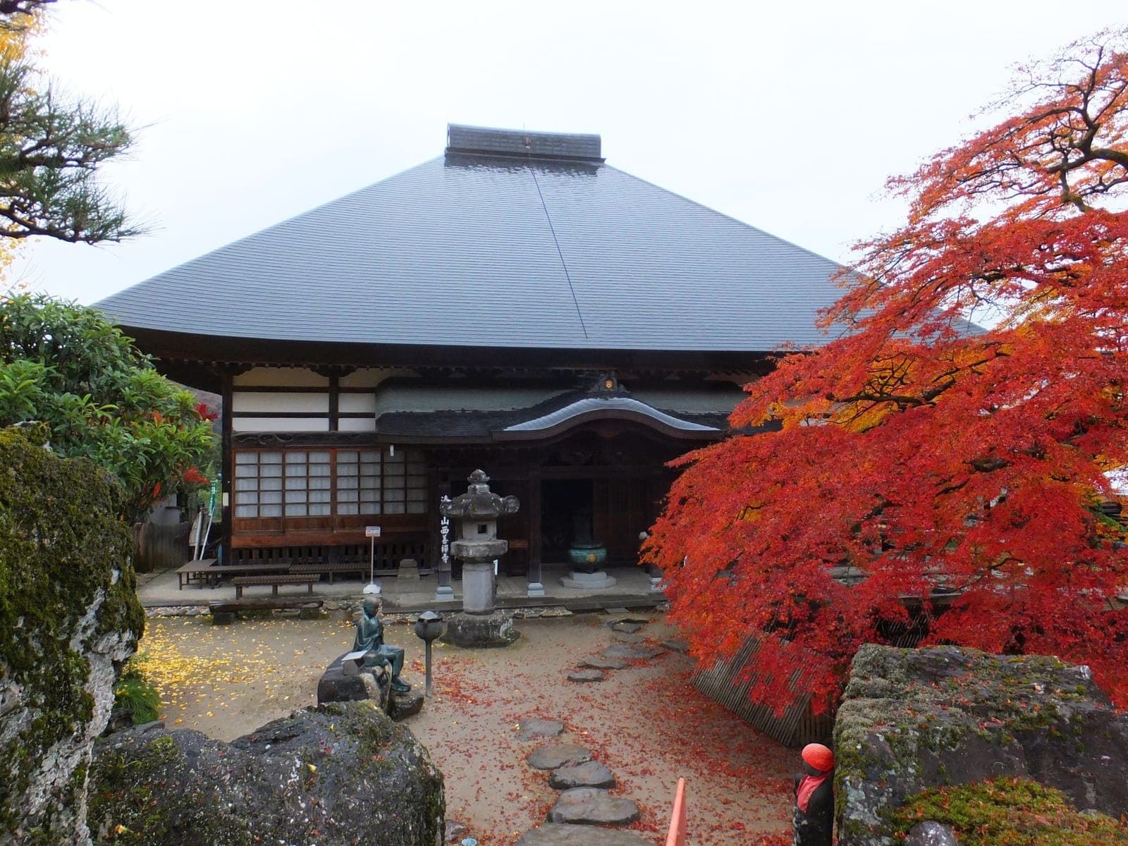 Saizen-ji temple exterior with a large dark roof, stone lantern, and red autumn maple leaves in the foreground.