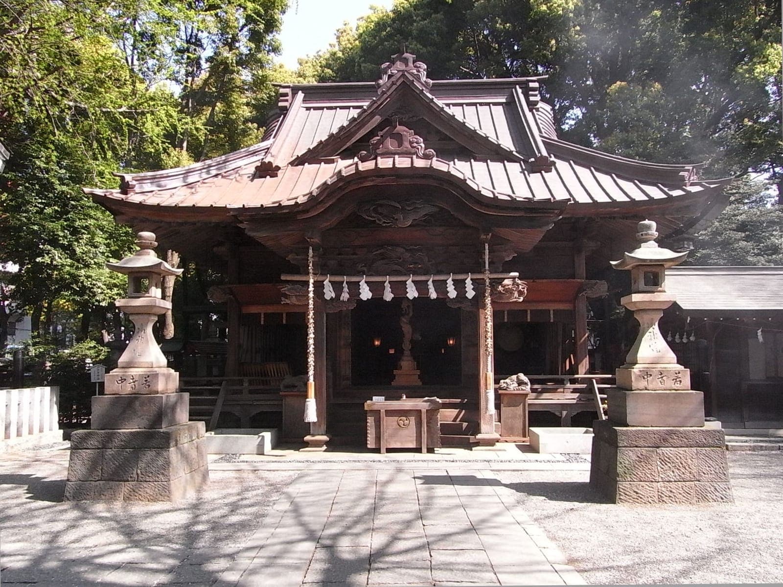 Exterior del santuario Tanashi con sala principal de madera, techo curvo, faroles de piedra y árboles.