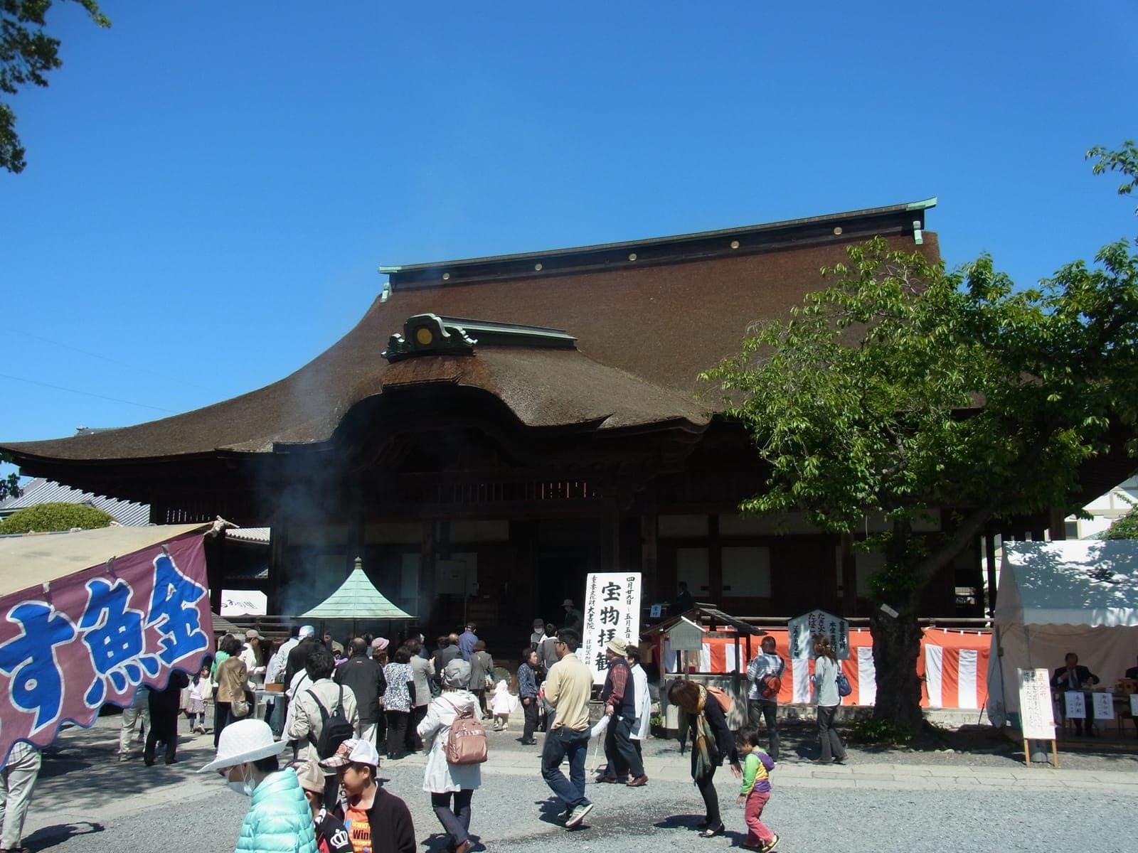 Extérieur du temple Mandara-ji, avec un grand toit courbé, une foule devant, des bannières et un arbre à droite.
