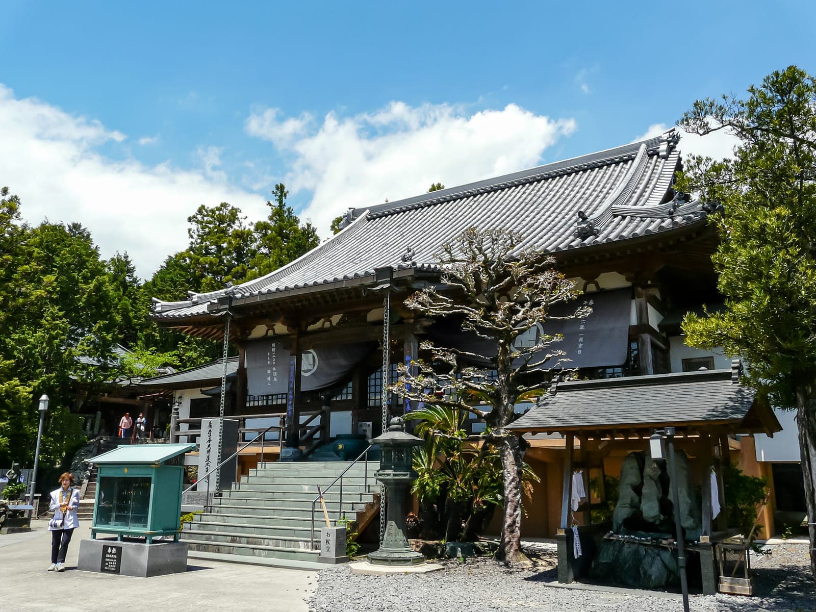 Juraku-ji temple exterior with a tiled roof, steps, lanterns, trees, and a small shrine basin in front.