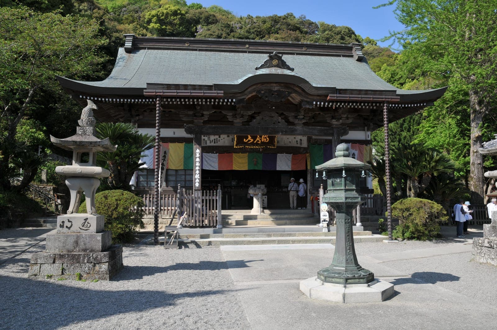 Außenansicht von Kirihata-ji mit breitem geschwungenem Dach, Steinlaternen und bunten Vorhängen, umgeben von grünen Bäumen.