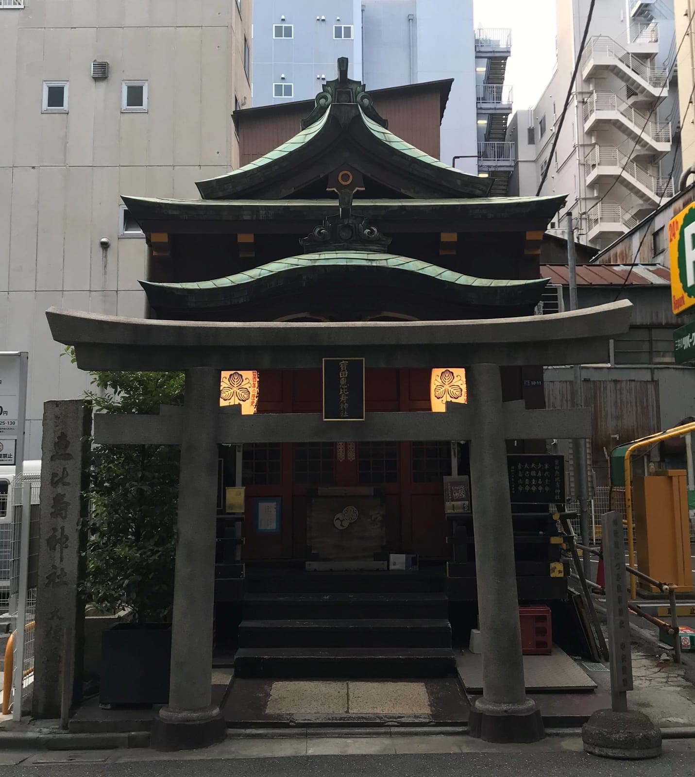 Entrada del santuario Takarada Ebisu con torii de piedra, escalones y techo verde, rodeada de edificios altos.