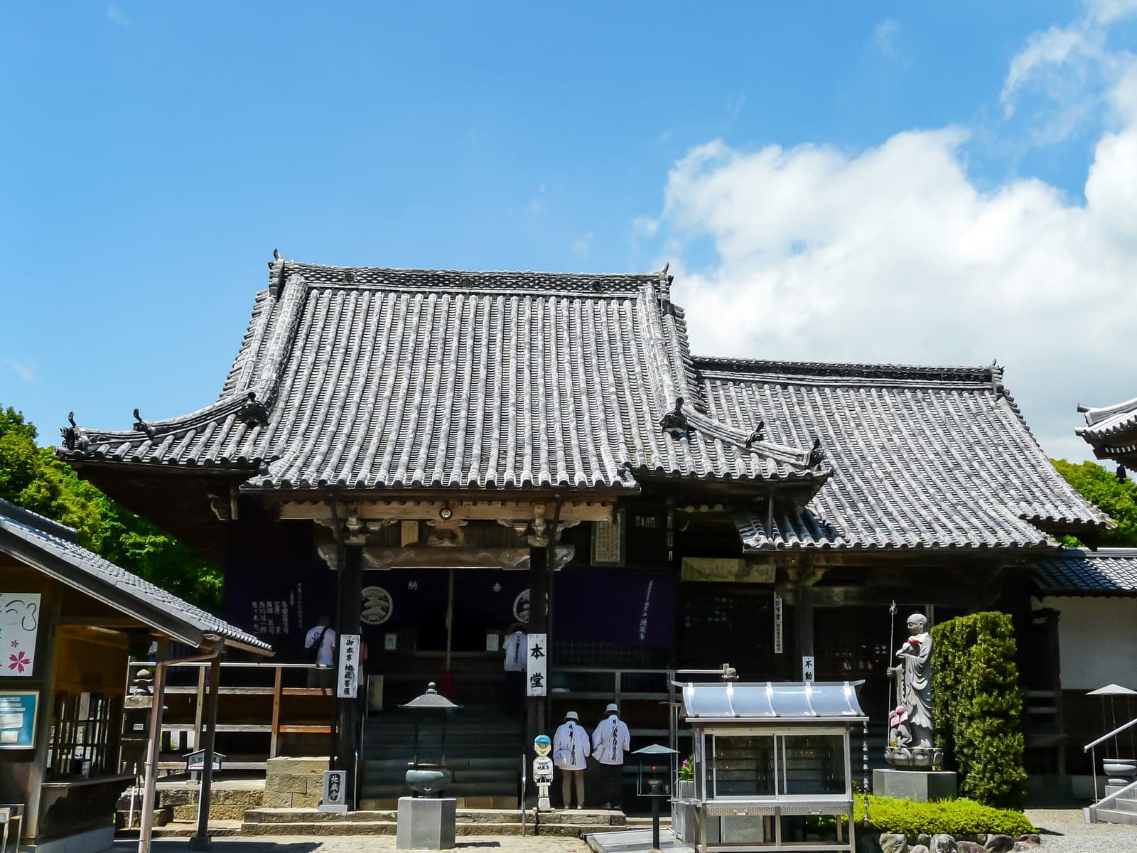 Jizo-ji temple exterior with a large tiled roof, entrance steps, and a statue beside the building under a blue sky.