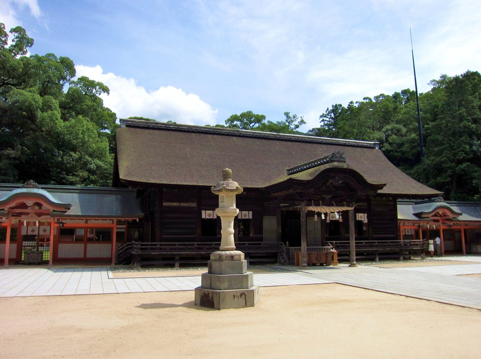 Oyamazumi Jinja exterior with a large thatched-roof shrine building, stone lantern in front, and trees behind.