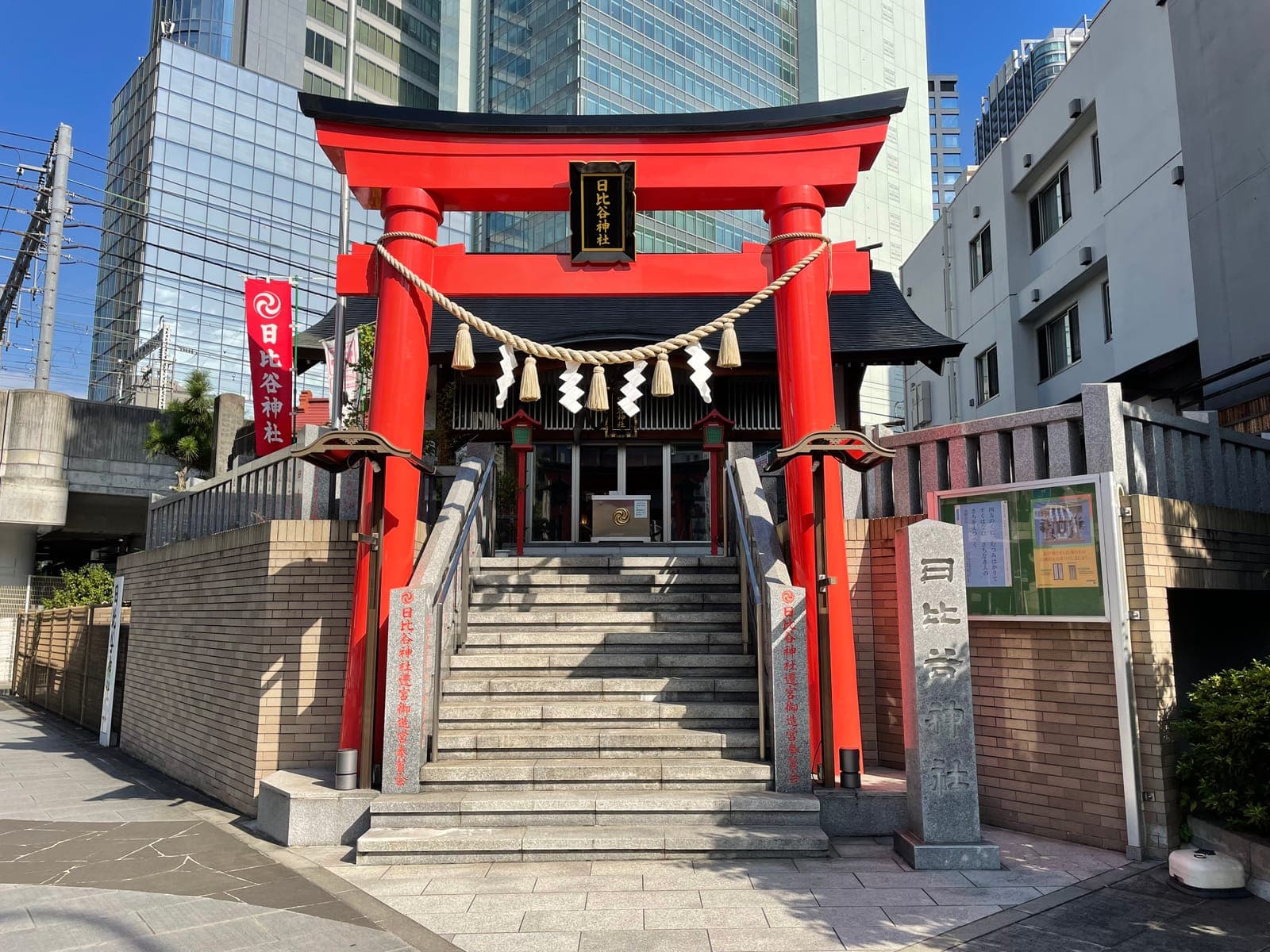 Entrada do santuário Hibiya com um torii vermelho vivo, escadaria de pedra e prédios modernos ao fundo.