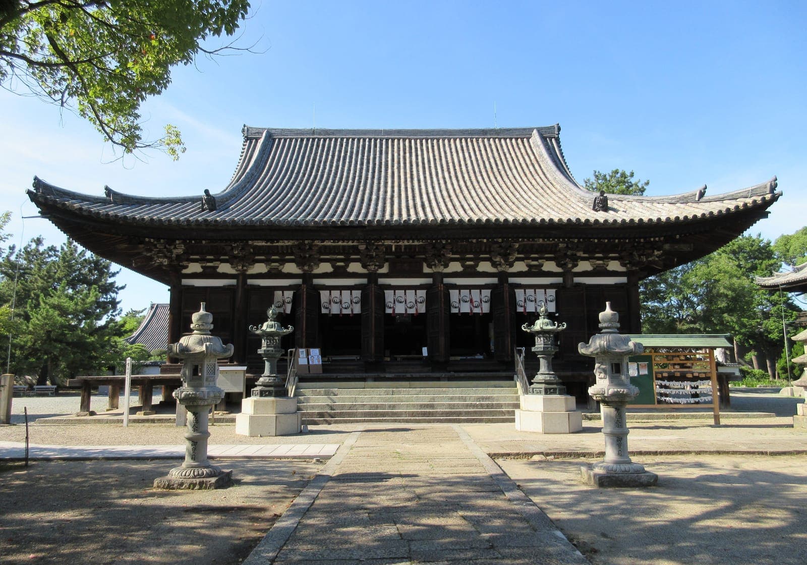 Salle principale du temple Kakurin-ji avec large toit en tuiles, lanternes de pierre et chemin menant aux marches sous un ciel bleu.