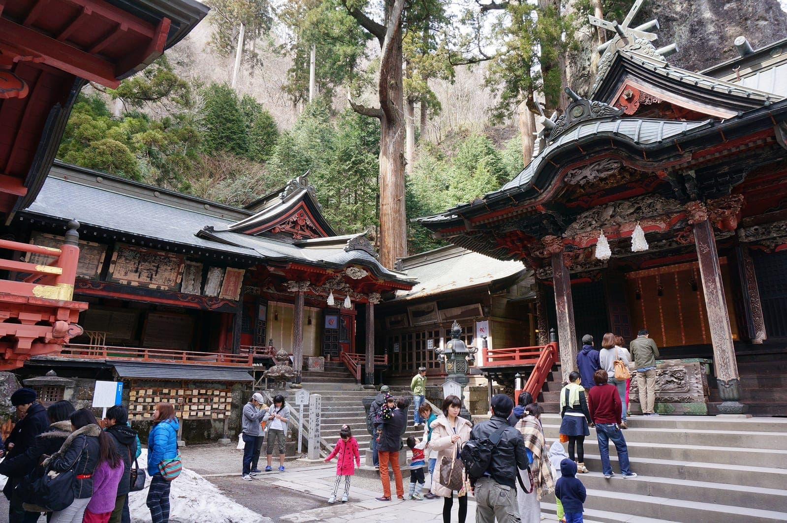 Patio exterior de Haruna Jinja con edificios del santuario rojos y negros ornamentados, escalones de piedra, árboles altos y visitantes.