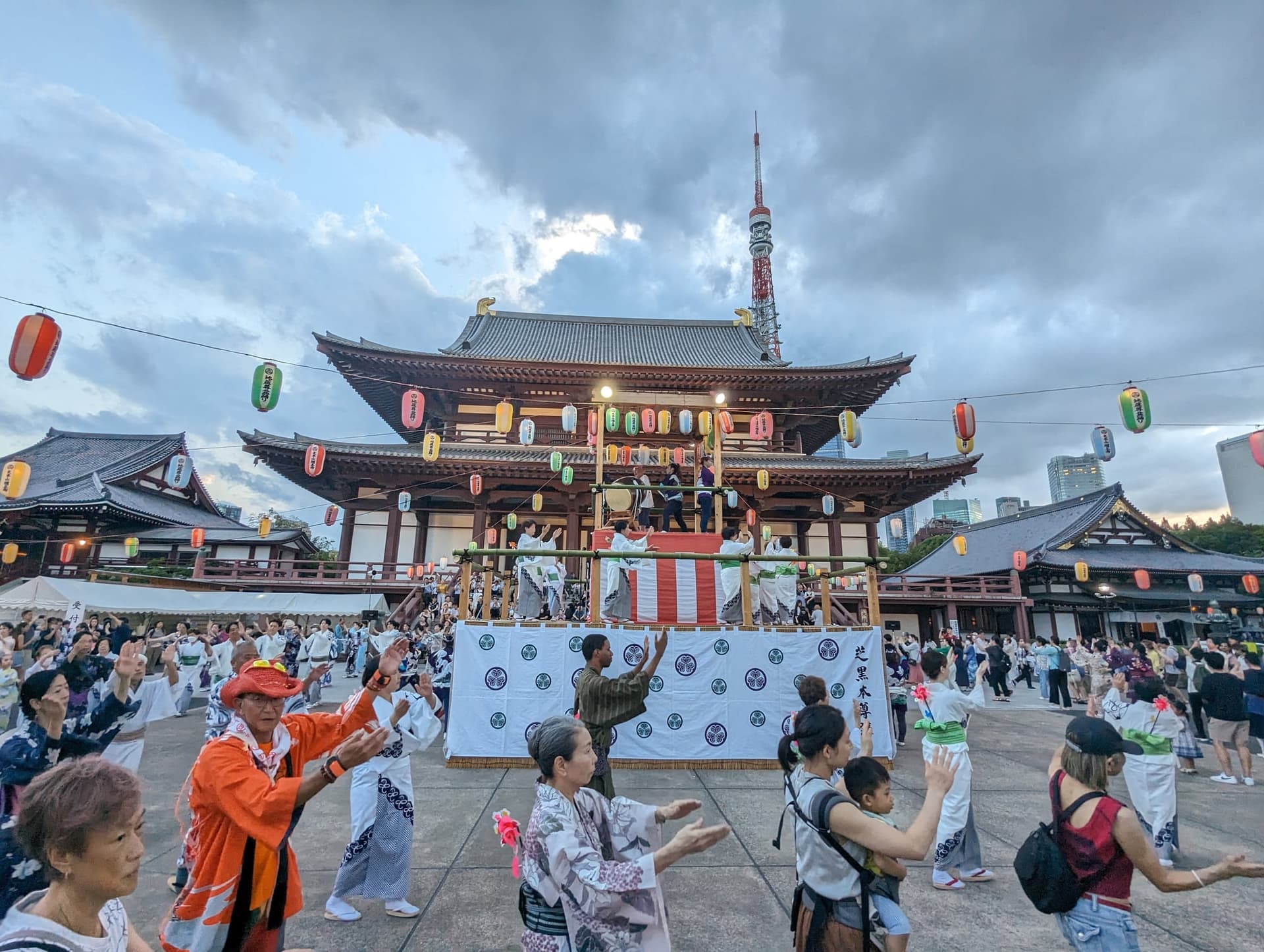Zojo-ji temple courtyard with lanterns strung overhead, people dancing in front of the main hall, and Tokyo Tower in the background.