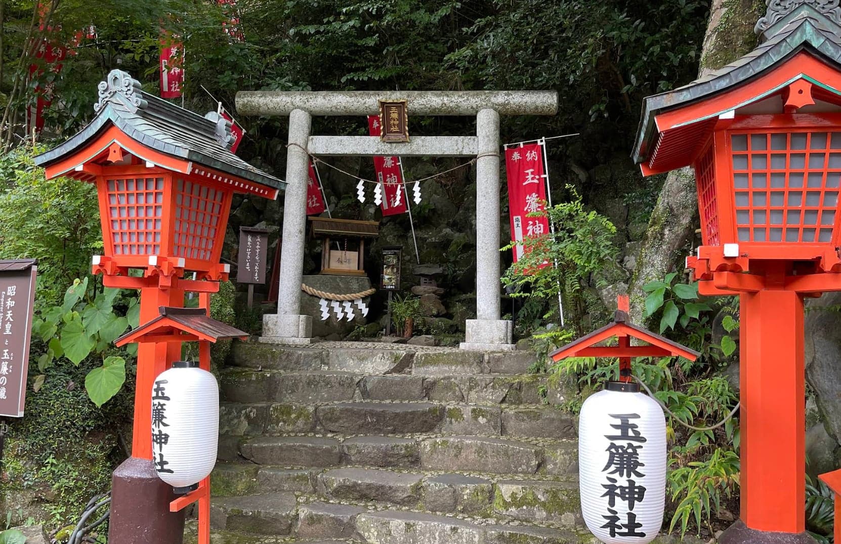 Torii de pedra e degraus para o santuário Tamasudare, emoldurados por lanternas e bandeiras vermelhas em área arborizada.