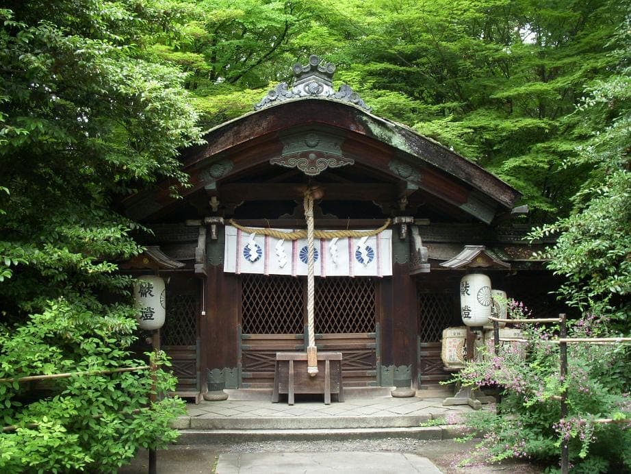 Exterior do templo Nashinoki com pequeno edifício de madeira, corda sagrada, lanternas e árvores verdes densas.