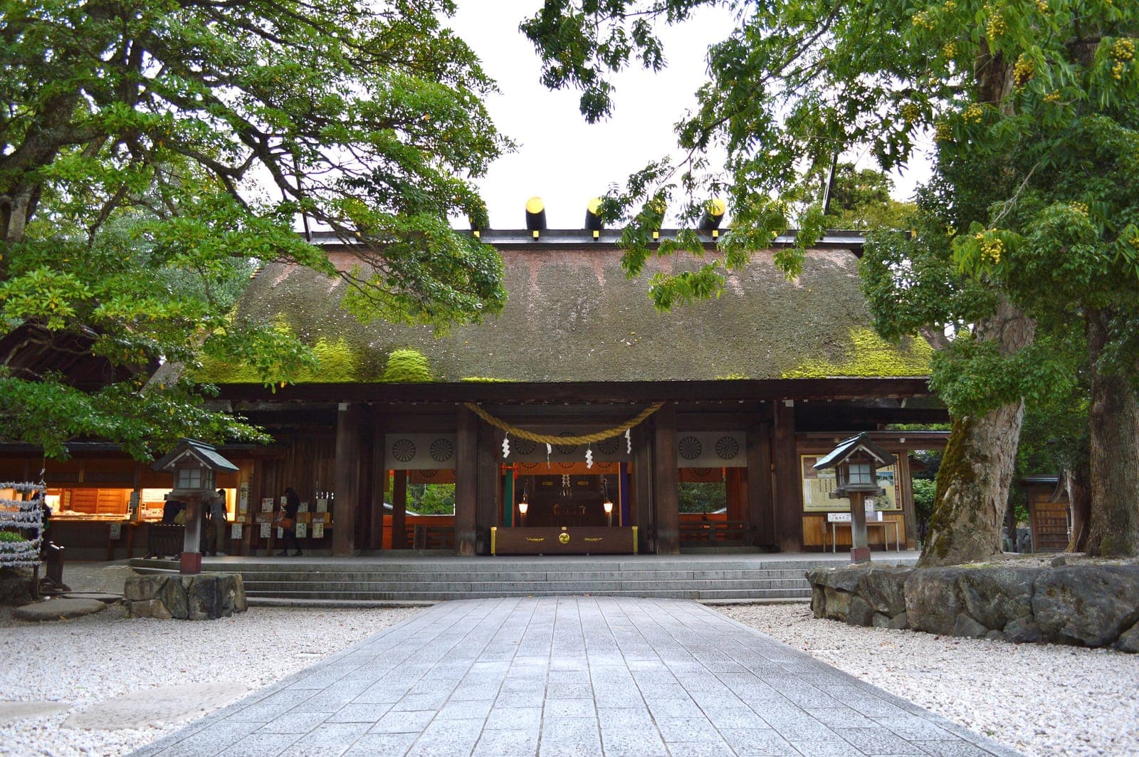 Exterior do santuário Motoise Kono, com telhado de palha, degraus de pedra e árvores emoldurando a entrada.
