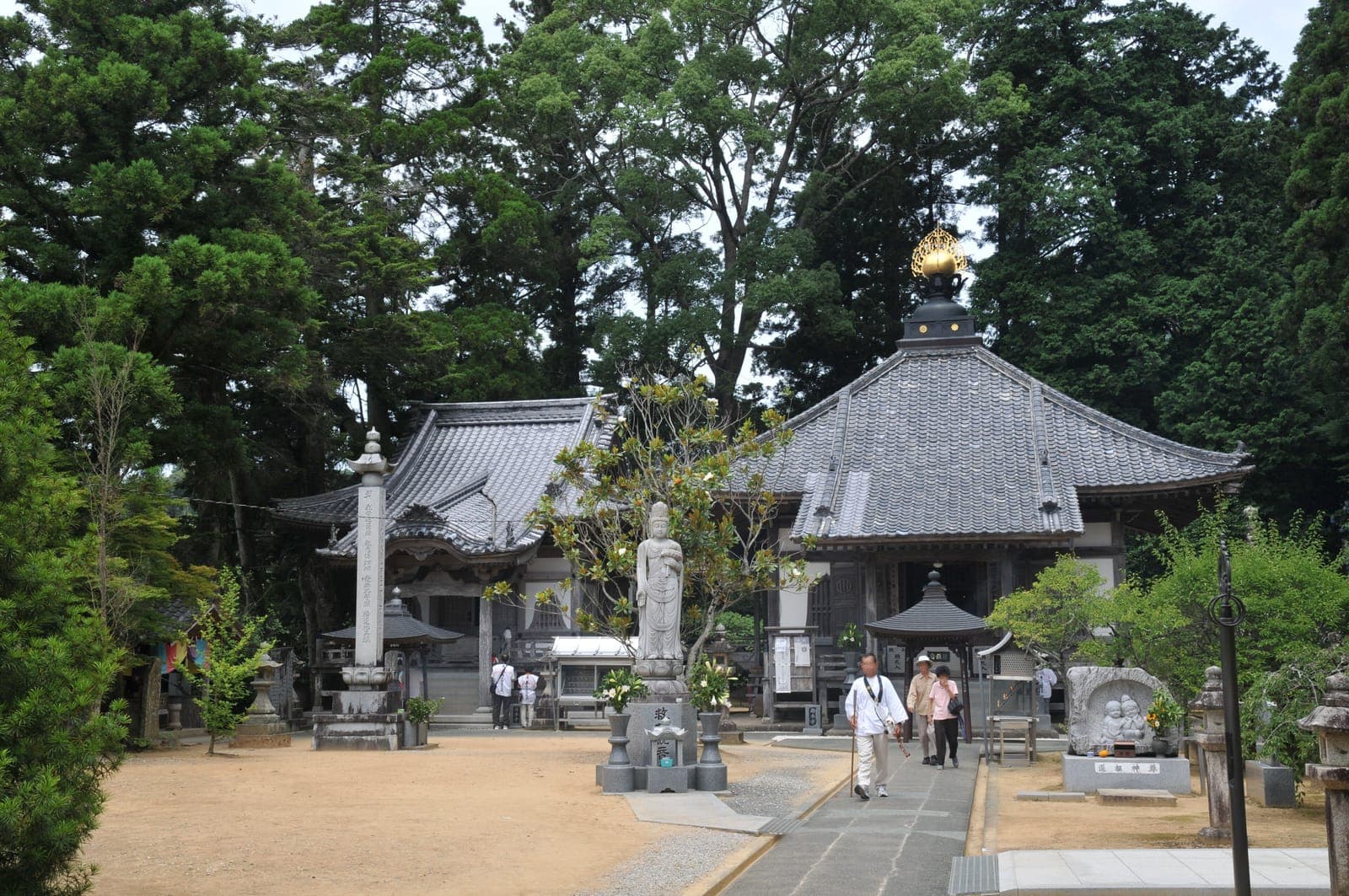 Butsumoku-ji temple courtyard with a tiled-roof hall, stone statues, and visitors among tall trees.