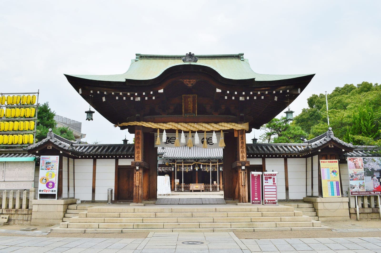Itatehyozu Jinja entrance gate and main hall with a curved green roof, stone steps, lanterns, and yellow paper lanterns at left.